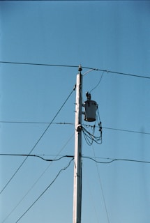 A wooden utility pole stands tall against a clear blue sky, with several wires extending from it. A transformer is mounted near the top of the pole, and multiple cables are wrapped around it.