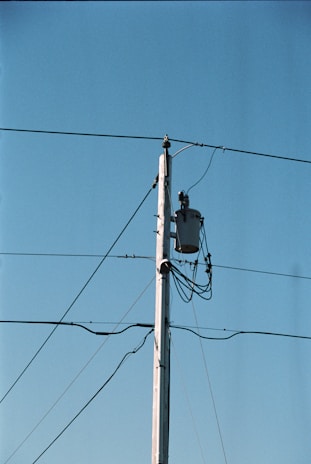 A wooden utility pole stands tall against a clear blue sky, with several wires extending from it. A transformer is mounted near the top of the pole, and multiple cables are wrapped around it.