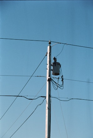 A wooden utility pole stands tall against a clear blue sky, with several wires extending from it. A transformer is mounted near the top of the pole, and multiple cables are wrapped around it.