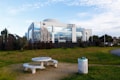 A modern building with reflective glass windows, displaying logos of Volkswagen and Audi. The foreground shows a picnic table and benches on a small patch of grass, next to a garbage can covered with graffiti. The area is surrounded by green lawns and small trees, under a partly cloudy sky.