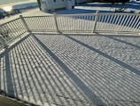 A snow-covered deck with a white railing casts long, linear shadows across the surface. The background shows a house and a snowy landscape under a clear sky, with sunlight reflecting off a distant body of water.