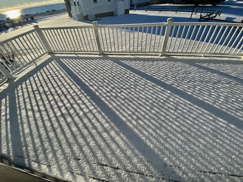 A snow-covered deck with a white railing casts long, linear shadows across the surface. The background shows a house and a snowy landscape under a clear sky, with sunlight reflecting off a distant body of water.