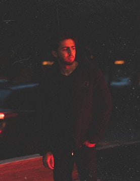 A person stands in a dimly lit environment with a red light casting a glow on their face and hand. The background is dark, enhancing the dramatic lighting effect.