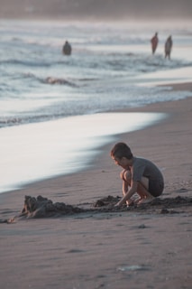 A child crouches on the beach, focused on building a sandcastle as the waves gently lap at the shore. The scene is bathed in the soft light of either dawn or dusk, creating a tranquil atmosphere. In the background, a few people walk along the water's edge, creating an impression of distance and space.