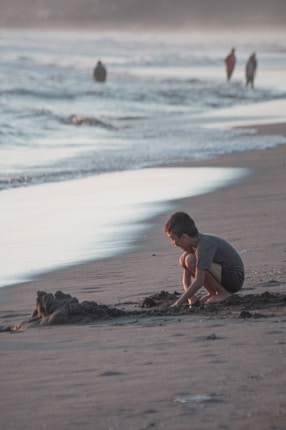 A child crouches on the beach, focused on building a sandcastle as the waves gently lap at the shore. The scene is bathed in the soft light of either dawn or dusk, creating a tranquil atmosphere. In the background, a few people walk along the water's edge, creating an impression of distance and space.
