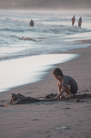 A child crouches on the beach, focused on building a sandcastle as the waves gently lap at the shore. The scene is bathed in the soft light of either dawn or dusk, creating a tranquil atmosphere. In the background, a few people walk along the water's edge, creating an impression of distance and space.