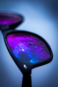 A close-up of a pair of sunglasses with lenses displaying a vibrant blend of purple and blue hues. Water droplets are scattered across the lens surface, creating a glossy, reflective appearance.