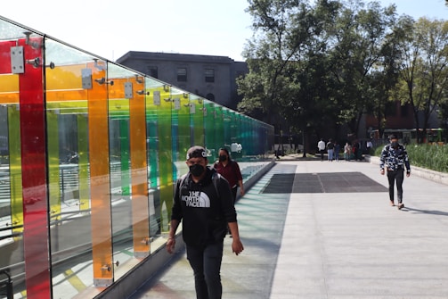 Several people wearing masks walk along a pathway next to a colorful, transparent wall made of vertical panels in red, orange, yellow, green, and blue. The setting is outdoors, with a large building in the background and trees providing shade along the sidewalk.