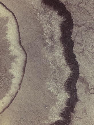 Abstract texture with wavy, dark lines and lighter, grainy areas. The surface appears rough, with a pattern resembling natural formations or erosion.