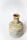 A ceramic bottle with a cylindrical body and narrow neck is positioned against a plain white background. The bottle has a rustic, handcrafted appearance with a muted color palette featuring earthy tones, such as shades of beige, light blue, and patches of orange-brown.