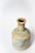A ceramic bottle with a cylindrical body and narrow neck is positioned against a plain white background. The bottle has a rustic, handcrafted appearance with a muted color palette featuring earthy tones, such as shades of beige, light blue, and patches of orange-brown.