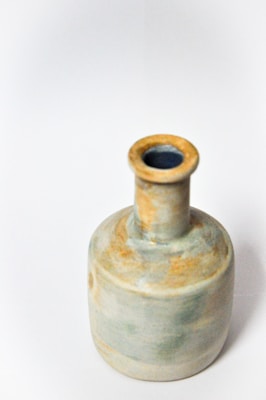 A ceramic bottle with a cylindrical body and narrow neck is positioned against a plain white background. The bottle has a rustic, handcrafted appearance with a muted color palette featuring earthy tones, such as shades of beige, light blue, and patches of orange-brown.