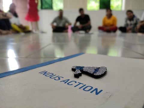 A group of people is seated on the floor in a circle, blurred in the background. In the foreground, there is a focus on a paper with the word 'INDUSACTION' written on it, and a decorative object made of rolled paper strips is placed atop it.