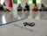 A group of people is seated on the floor in a circle, blurred in the background. In the foreground, there is a focus on a paper with the word 'INDUSACTION' written on it, and a decorative object made of rolled paper strips is placed atop it.