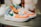 A pair of colorful athletic sneakers featuring a combination of vivid orange, green, blue, and beige panels. The shoes are displayed on a rounded surface, and have a distinct logo on the side. A tag is attached to one sneaker.