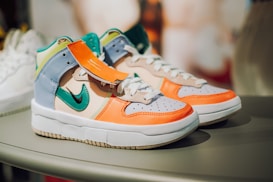 A pair of colorful athletic sneakers featuring a combination of vivid orange, green, blue, and beige panels. The shoes are displayed on a rounded surface, and have a distinct logo on the side. A tag is attached to one sneaker.