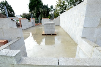 An unfinished building with concrete block walls under construction. The floor appears to be made of concrete and there is a stack of concrete blocks placed on a wooden pallet in the center. The area is open to the sky with some trees visible in the background.