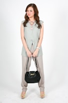 A woman with long, wavy brown hair is standing against a plain white background. She is dressed in a light green sleeveless top and loose beige pants. She is holding a black handbag with both hands in front of her. She is wearing a statement necklace with colorful gemstones, a watch on her left wrist, and yellow high-heeled sandals.