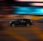 A car moving swiftly with a motion blur effect in the background, showcasing bright streaks of light in various colors, suggesting a cityscape or urban environment at night.