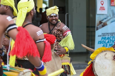 A group of people wearing traditional, colorful garments are playing drums. Their attire includes leopard print patterns and bright yellow and red tassels. They have face paint and are performing energetically in an outdoor setting.