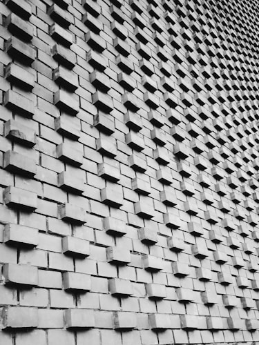 A wall constructed with an array of protruding bricks arranged in a uniform, repetitive pattern. The bricks create a textured surface with shadows enhancing the three-dimensional effect. The perspective adds a sense of depth, as the wall appears to curve slightly.