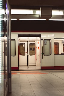 An open metro train door is visible, with a view of the interior showing empty seats. The station platform appears deserted, creating a sense of stillness. Overhead lighting casts a warm glow, and reflections are seen on the tiled floor.