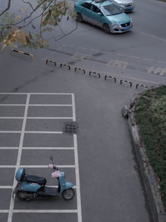 A parking area with painted white lines featuring an unoccupied blue scooter positioned in one of the spaces. Above the parking area, a road is visible with a teal car driving through. On the side of the road, some greenery and leaves are present, possibly a small garden or verge. The branches of a tree with green and yellow leaves frame the top edge of the scene.