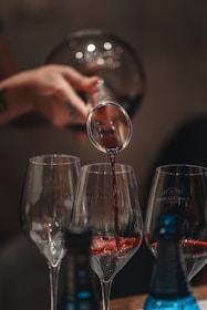 Close-up of hands pouring deep red wine into elegant glasses amid terracotta and olive green tones.