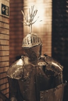 A suit of medieval armor stands on display in a dimly lit room. The armor is metallic, featuring intricate patterns etched into the helmet and chest plate. A decorative plume adorns the top of the helmet. The background consists of brick walls and a partially visible directional sign.