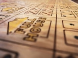 A close-up view of a wooden boardgame surface with engraved patterns, including the words 'FULL HOUSE' and various sections marked with stylized dots resembling dice. The focus is shallow, highlighting the text and some dot patterns while the rest of the board fades softly into the background.