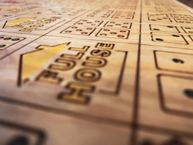 A close-up view of a wooden boardgame surface with engraved patterns, including the words 'FULL HOUSE' and various sections marked with stylized dots resembling dice. The focus is shallow, highlighting the text and some dot patterns while the rest of the board fades softly into the background.