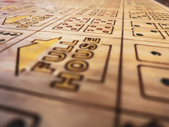 A close-up view of a wooden boardgame surface with engraved patterns, including the words 'FULL HOUSE' and various sections marked with stylized dots resembling dice. The focus is shallow, highlighting the text and some dot patterns while the rest of the board fades softly into the background.