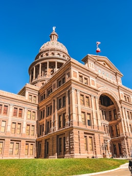 A large historic building with a prominent dome, featuring classical architecture and numerous windows. The facade is made of stone with intricate details and symmetrical design. A flag flutters on top of the dome against a clear blue sky.