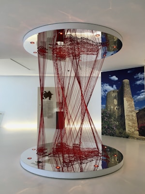 An intricate art installation featuring numerous red threads strung between two mirrored circular surfaces, creating a visually compelling and elaborate pattern. The surrounding environment appears to be a modern gallery space with white walls. In the background, there is a large photograph showcasing a historic stone tower with a blue sky and some clouds.