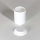 A white cylindrical container with a separate lid, both made of a smooth, opaque material. The container and lid are positioned on a light gray surface, casting soft shadows.