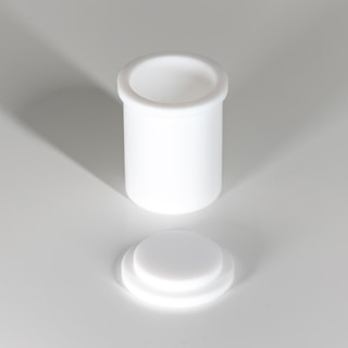 A white cylindrical container with a separate lid, both made of a smooth, opaque material. The container and lid are positioned on a light gray surface, casting soft shadows.