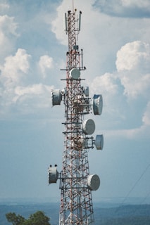 A tall communication tower with multiple antenna dishes and panels, set against a backdrop of a clear sky with scattered clouds. The structure is composed of metal lattice and the surrounding landscape features distant hills and vegetation.