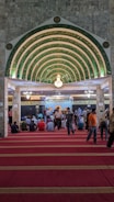 An ornate, Islamic architectural interior with a series of green and gold archways featuring intricate Arabic calligraphy. A chandelier hangs from the ceiling. People are seated on a red carpet, some are standing, indicating a gathering or prayer session.