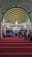 An ornate, Islamic architectural interior with a series of green and gold archways featuring intricate Arabic calligraphy. A chandelier hangs from the ceiling. People are seated on a red carpet, some are standing, indicating a gathering or prayer session.