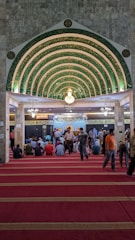 An ornate, Islamic architectural interior with a series of green and gold archways featuring intricate Arabic calligraphy. A chandelier hangs from the ceiling. People are seated on a red carpet, some are standing, indicating a gathering or prayer session.