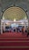 An ornate, Islamic architectural interior with a series of green and gold archways featuring intricate Arabic calligraphy. A chandelier hangs from the ceiling. People are seated on a red carpet, some are standing, indicating a gathering or prayer session.