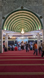 An ornate, Islamic architectural interior with a series of green and gold archways featuring intricate Arabic calligraphy. A chandelier hangs from the ceiling. People are seated on a red carpet, some are standing, indicating a gathering or prayer session.