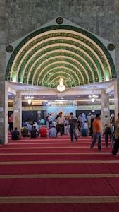 An ornate, Islamic architectural interior with a series of green and gold archways featuring intricate Arabic calligraphy. A chandelier hangs from the ceiling. People are seated on a red carpet, some are standing, indicating a gathering or prayer session.