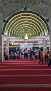 An ornate, Islamic architectural interior with a series of green and gold archways featuring intricate Arabic calligraphy. A chandelier hangs from the ceiling. People are seated on a red carpet, some are standing, indicating a gathering or prayer session.