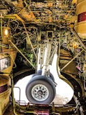 The intricate interior of an aircraft landing gear compartment, featuring a complex network of wires, pipes, and mechanical components surrounding a large wheel. The lighting and metallic surfaces create a technical and industrial feel.