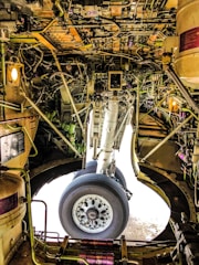 The intricate interior of an aircraft landing gear compartment, featuring a complex network of wires, pipes, and mechanical components surrounding a large wheel. The lighting and metallic surfaces create a technical and industrial feel.