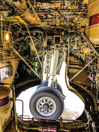 The intricate interior of an aircraft landing gear compartment, featuring a complex network of wires, pipes, and mechanical components surrounding a large wheel. The lighting and metallic surfaces create a technical and industrial feel.