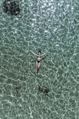 A person is floating peacefully on their back in clear, turquoise water. Sunlight creates intricate patterns on the surface of the water, giving it a shimmering, textured appearance. The person is wearing a black swimsuit, and the scene exudes a sense of tranquility and leisure.