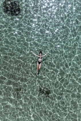 A person is floating peacefully on their back in clear, turquoise water. Sunlight creates intricate patterns on the surface of the water, giving it a shimmering, textured appearance. The person is wearing a black swimsuit, and the scene exudes a sense of tranquility and leisure.