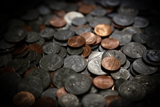 A close-up of a scattered pile of coins, featuring various denominations and colors, primarily silver and copper. The lighting creates a dramatic contrast, highlighting the reflective surfaces of the coins.